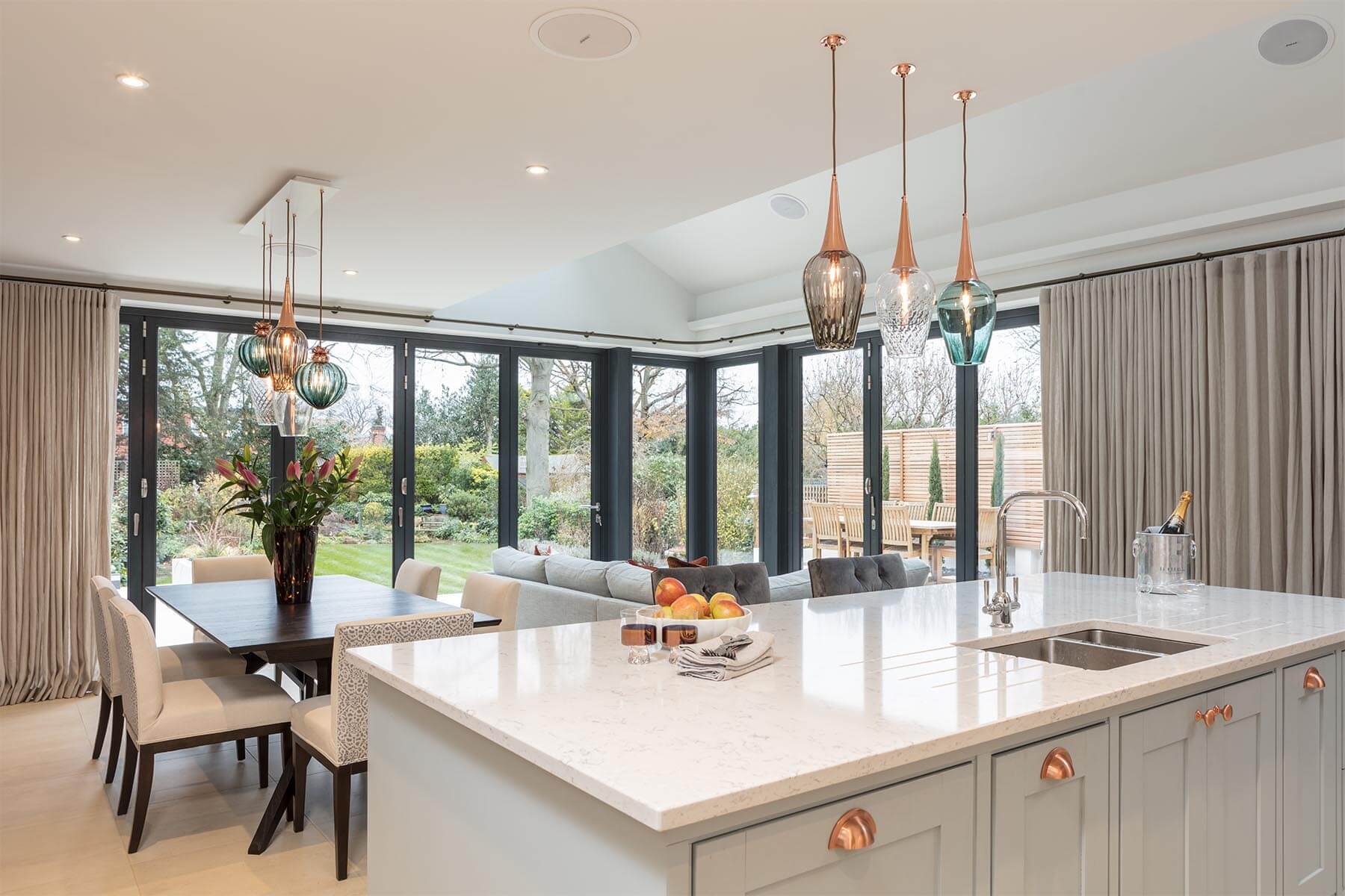 A modern kitchen features a marble countertop with a sink, surrounded by elegant lighting, a dining table with chairs, and views of a garden through large glass doors.