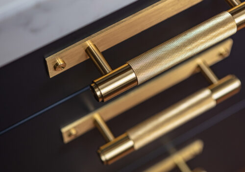 A polished brass cabinet handle is prominently displayed, featuring a textured grip. It's positioned against a dark cabinet surface, suggesting a contemporary design in a kitchen or furniture setting.