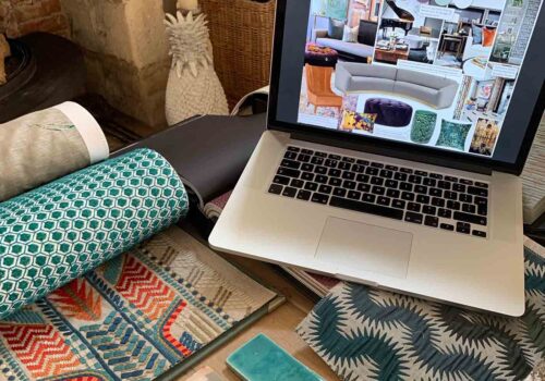 A laptop displays design inspiration while colorful fabric swatches and a tile sample are spread around. The setting includes natural light and textured surfaces, suggesting a creative workspace.