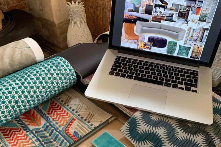 A laptop displays design inspiration while colorful fabric swatches and a tile sample are spread around. The setting includes natural light and textured surfaces, suggesting a creative workspace.