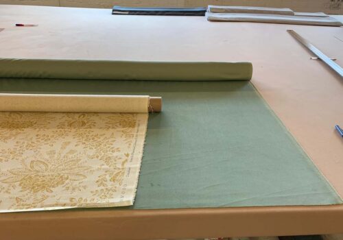 A roll of patterned fabric lies on a working table, partially unrolled next to a solid-colored green fabric. Various fabric rolls and tools are visible in the background.