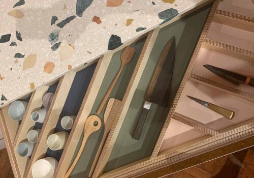 Wooden drawers display various kitchen utensils, including knives and stirring spoons. The countertop features a speckled design, adding a colorful, textured background to the organized layout of tools.