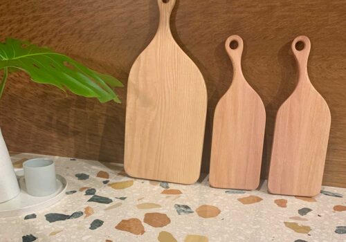 Three wooden serving boards of varying sizes stand upright against a brown wall. A green leaf plant and a small cup on a speckled countertop complete the kitchen setting.