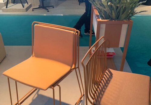 Two modern chairs in soft orange and beige are positioned on a light-colored floor. A planter with greenery is nearby, and a teal carpet adds depth to the setting.