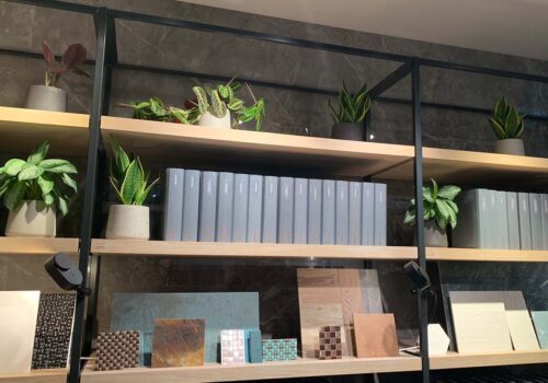 A shelf displays various potted plants alongside stacked gray binders and decorative samples, including tiles and wood pieces, against a marble wall in a modern indoor setting.