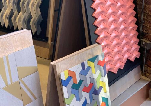 Colorful wall tiles in varying geometric patterns are displayed upright. They include textured designs in pink, gray, and multicolored shapes, set against a modern interior backdrop.