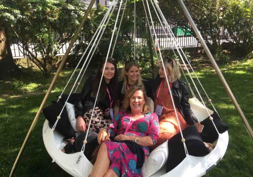 A large hanging swing chair holds four women, smiling and posing together. They are in a green garden area surrounded by trees, enjoying a sunny day.