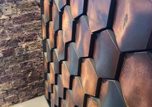 A hexagonal patterned wall surface showcases a blend of earthy tones, with textured elements catching light. Behind it, a rustic brick wall adds depth to the setting.