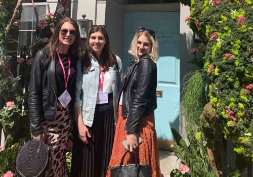 Three women stand together in front of a light blue door adorned with greenery and flowers. They are smiling, wearing stylish outfits, and holding handbags. The sun is shining.