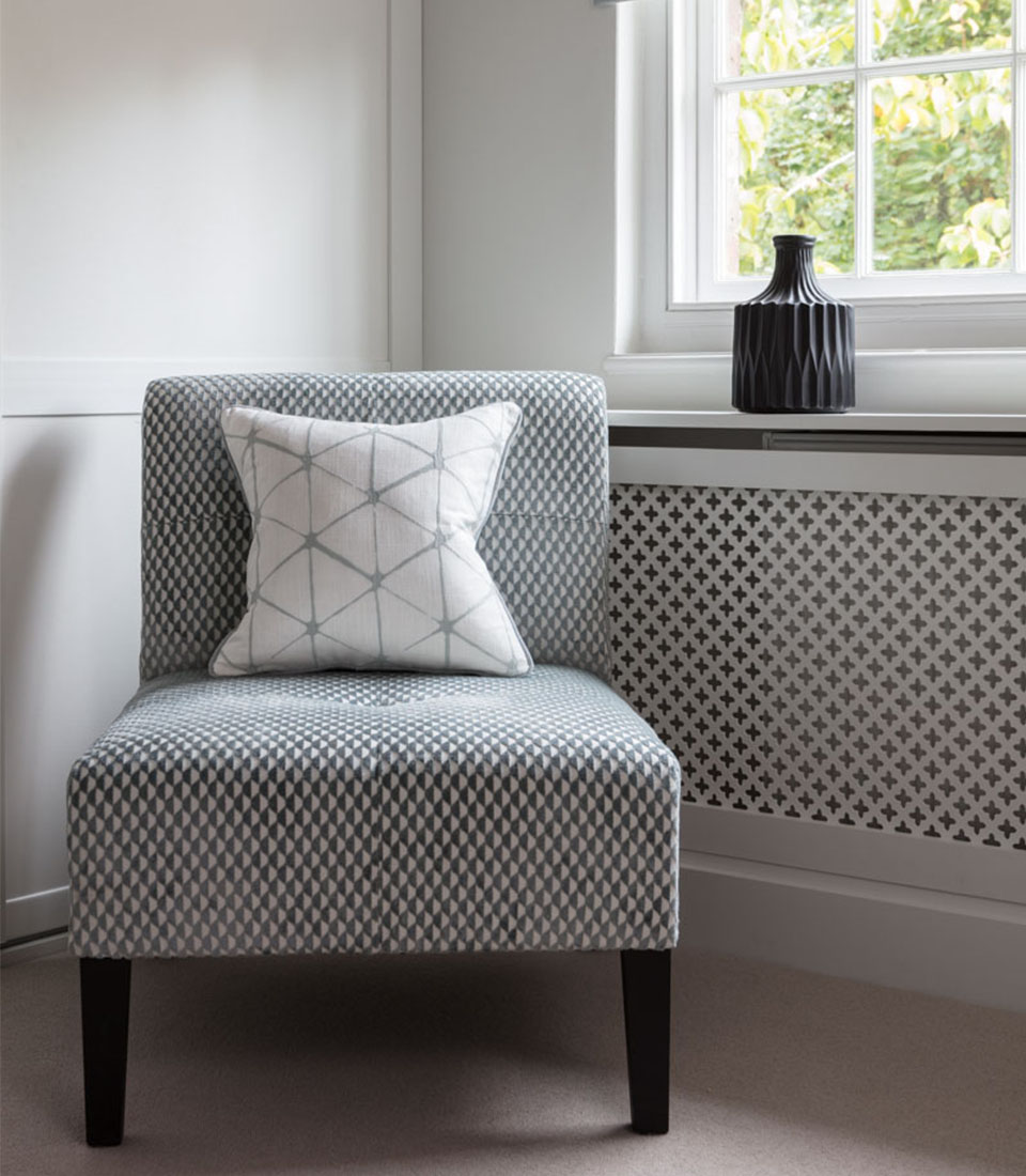 A patterned armchair with a decorative pillow sits beside a window, against a wall featuring intricate geometric designs. Light filters through, creating a calm, inviting atmosphere.