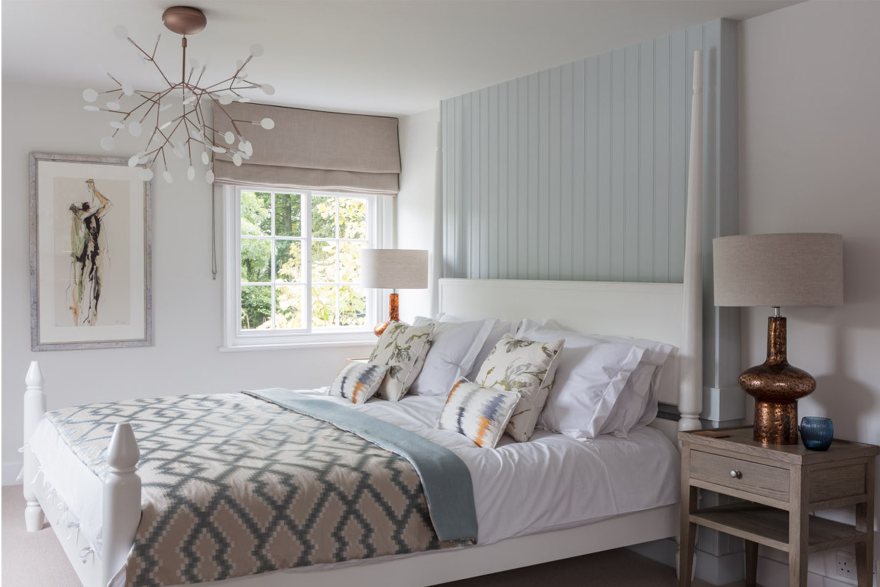 A double bed with decorative pillows is neatly arranged in a bright bedroom. A stylish chandelier hangs above, while natural light filters through a window beside a bedside table with a lamp.
