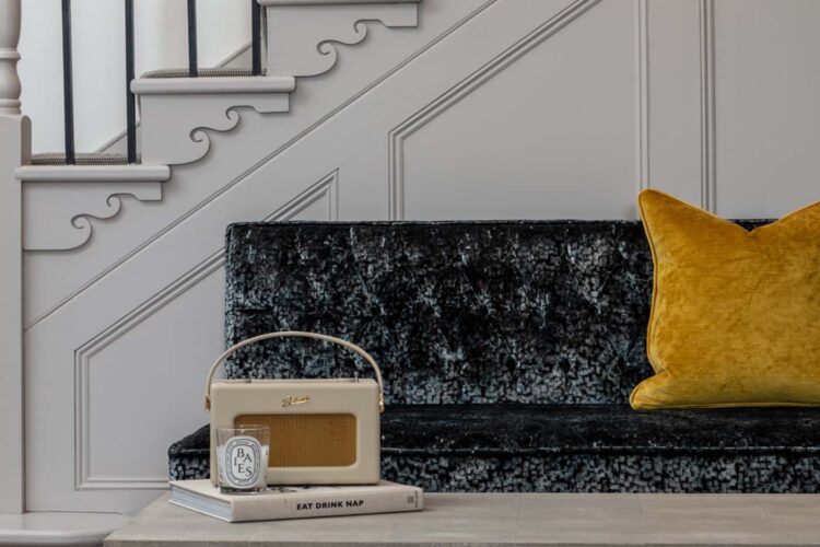 A plush black sofa with a yellow pillow features a vintage radio and a candle on stacked books. The background includes elegant stairs with decorative railings and molded walls.