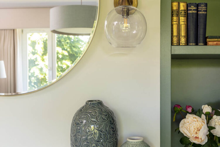 A decorative mirror reflects a clear glass wall light above two intricate ceramic vases on a mantel. A nearby bookshelf holds various books, and a bouquet of peonies adds color.