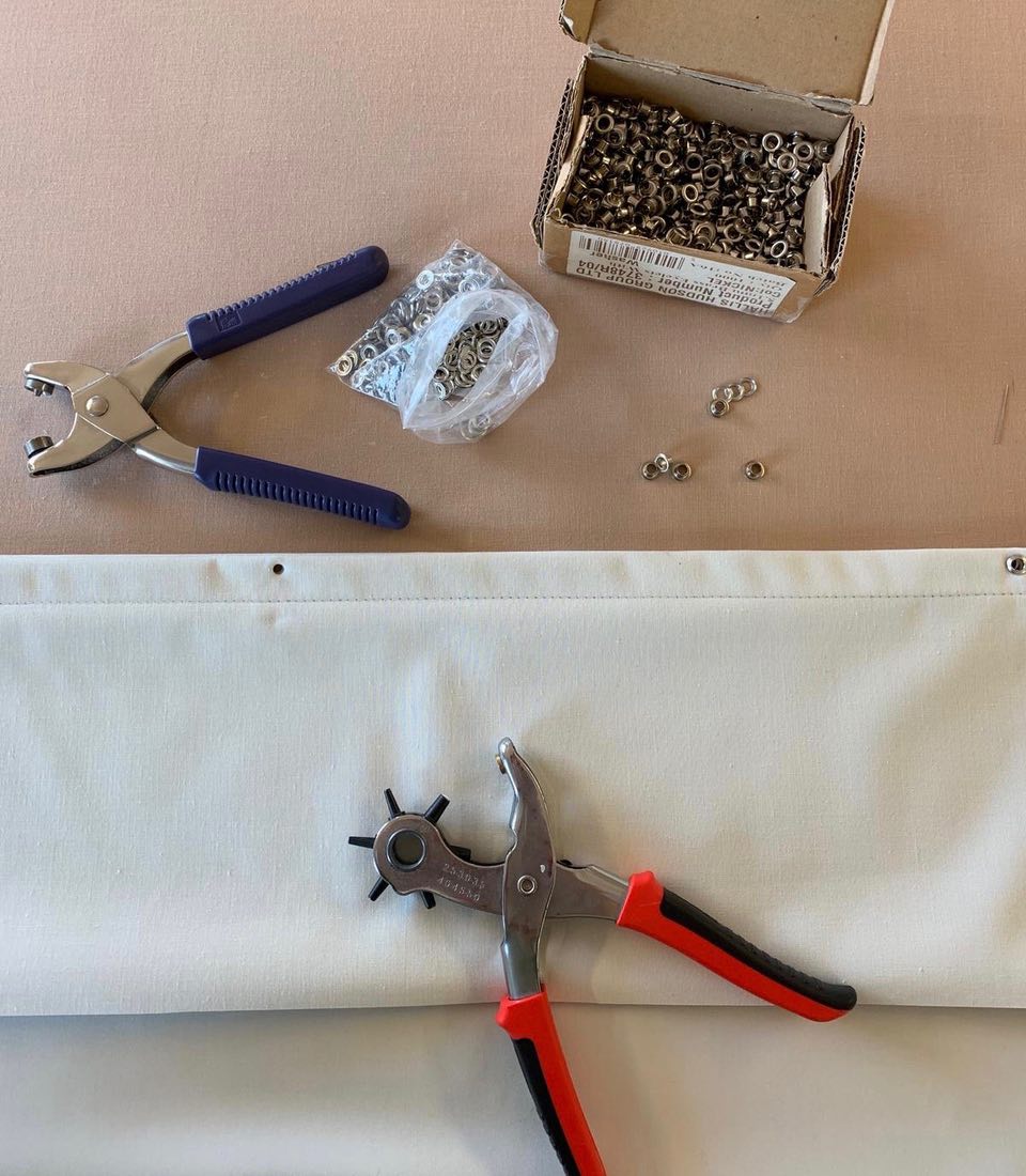 A pair of grommet pliers is positioned next to a piece of fabric with a hole, surrounded by a box and a bag of metal grommets on a beige surface.