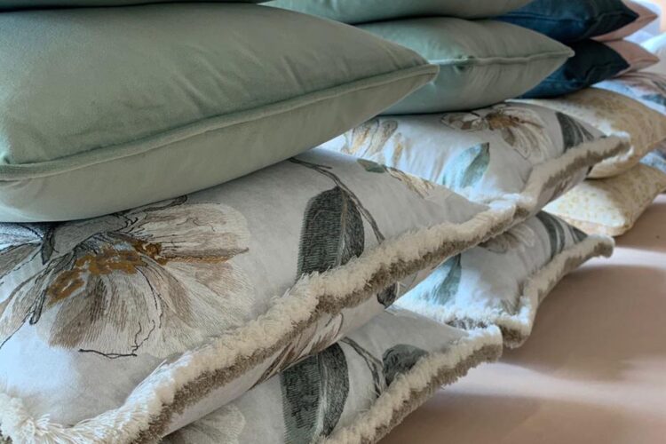 A stack of decorative cushions is arranged neatly. The top cushions are solid green, while lower ones feature floral patterns with fringe edges. They rest on a light-colored surface.