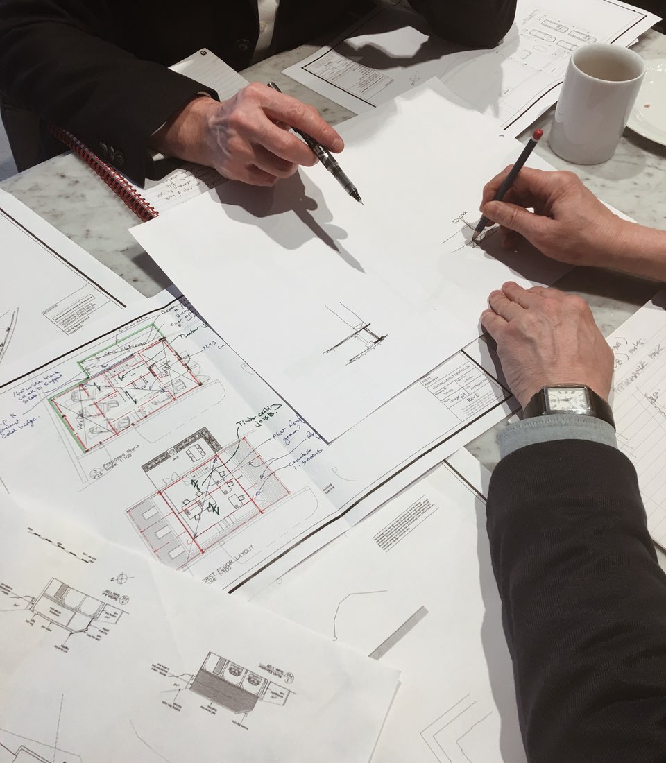 Two hands are engaged in sketching architectural plans on papers spread across a table, surrounded by notebooks, a cup, and a plate, indicating collaboration in a design discussion.