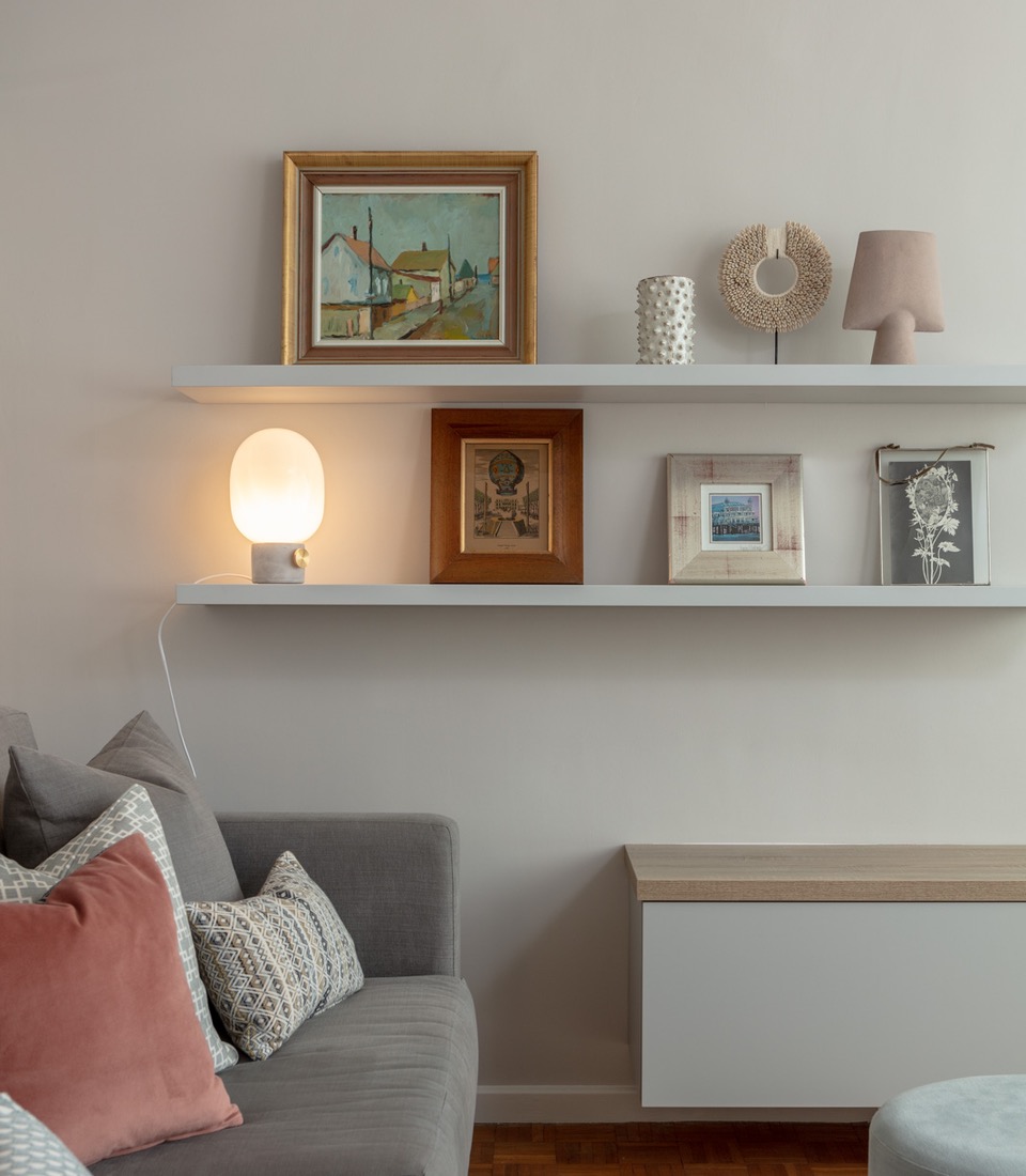 A modern living space features a gray sofa adorned with decorative pillows. Above, a white shelf holds paintings and decorative objects, including a lamp casting a warm glow.