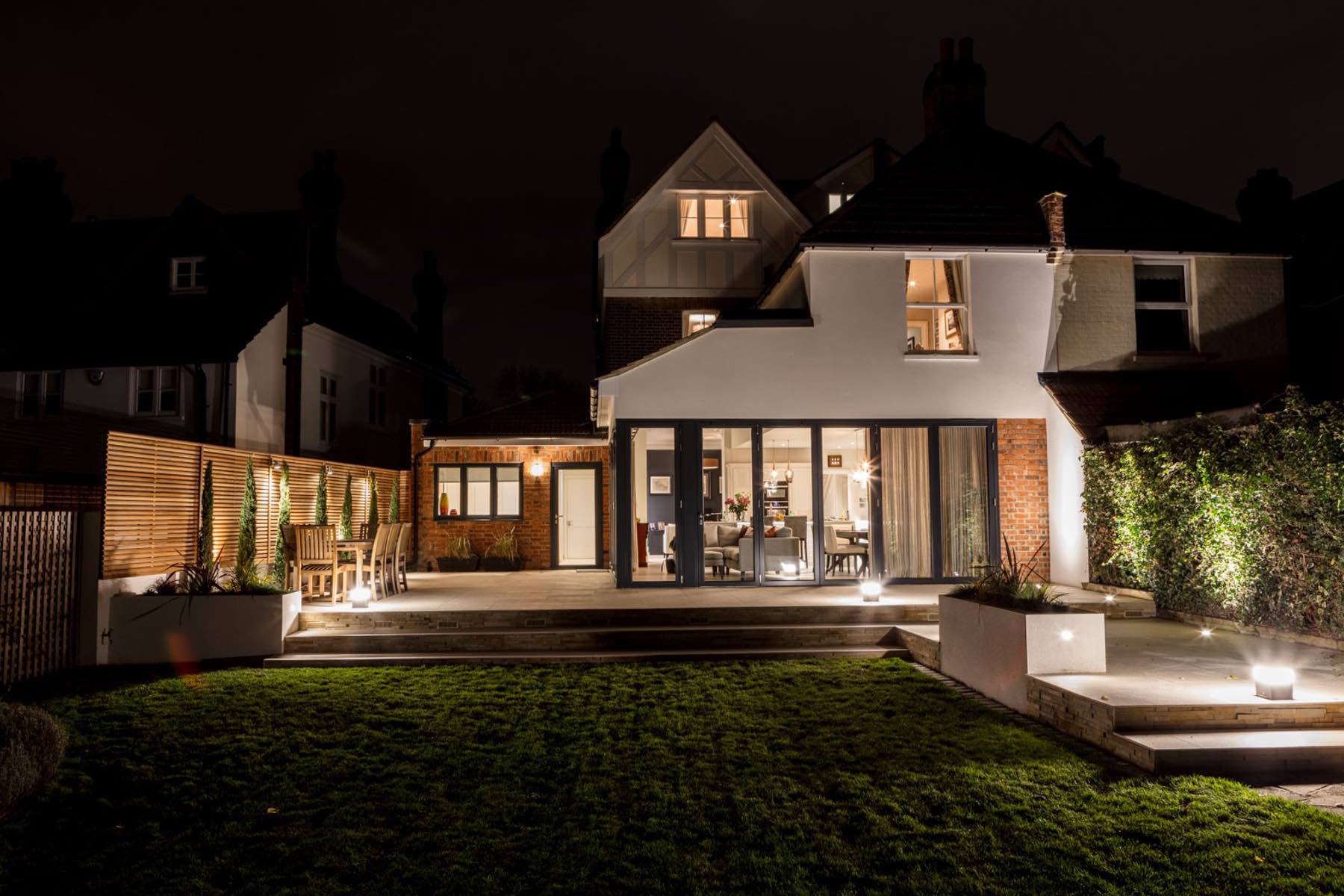 A well-lit modern home features a patio with outdoor furniture and greenery, illuminated by soft lights, surrounded by dark skies and neighboring houses at night.