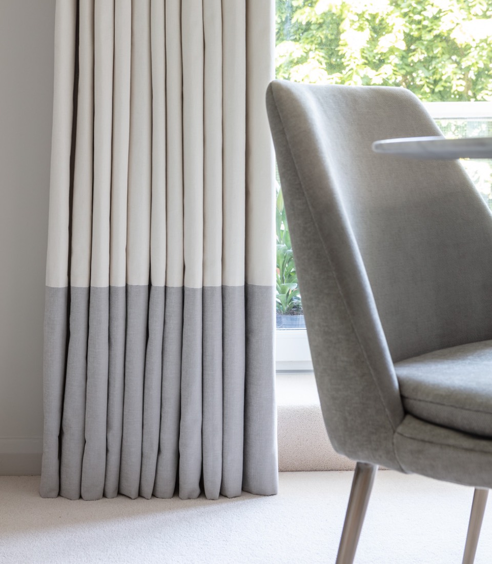 White and gray striped curtains hang beside a window, gently pooling on a beige carpet. A modern gray chair is positioned nearby, suggesting a calm, contemporary living space.