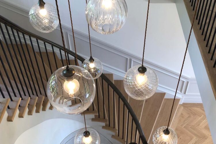 Hanging glass light fixtures illuminate a spiral staircase, casting soft light in a modern, minimalist interior. The stair treads are wooden, contrasted by a dark railing and white walls.