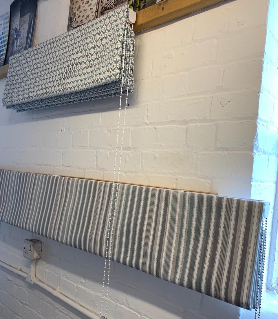 Roller blinds with leaf and striped patterns are displayed on a white brick wall. One blind is rolled up, while another is partially rolled down, showing the fabric designs. A window is visible in the background.