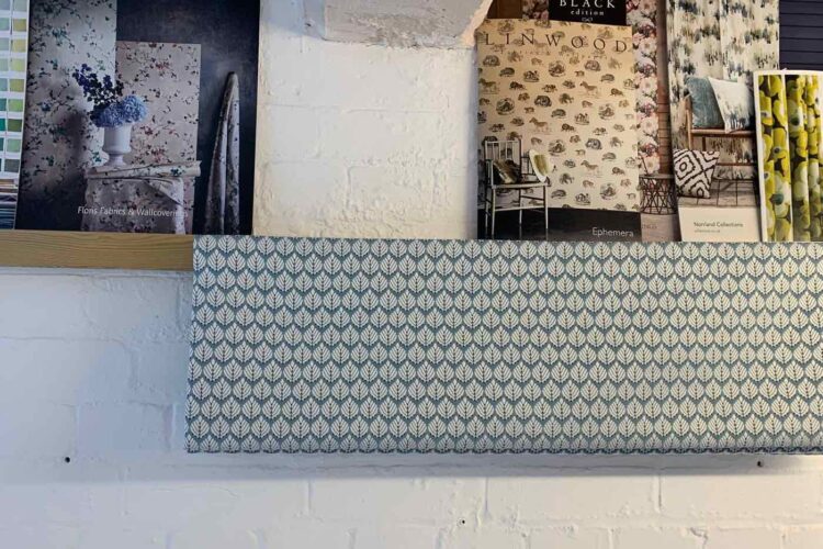 A shelf displays several fabric sample books, showcasing various floral and patterned designs, with a patterned fabric swatch hanging below, all set against a white brick wall.