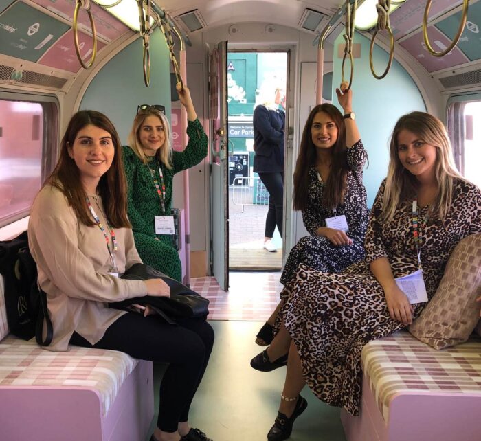 Four women are seated in a colorful, vintage-style train interior, smiling and posing for a photo. They are dressed in varied patterns, with a view of an open train door behind them.