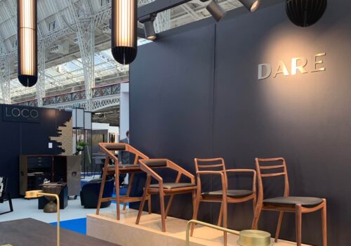 Four stylish wooden chairs with black cushions are arranged against a dark wall labeled “DARE,” within a modern exhibition space featuring overhead lighting and minimalistic decor.