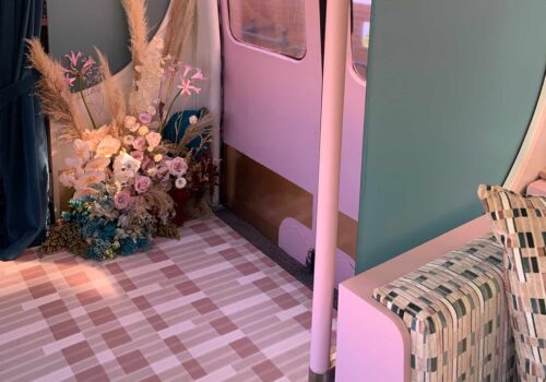 A floral arrangement in soft hues is positioned on a patterned floor near a pink and green wall. A cozy, patterned couch and curtains add a warm atmosphere to the inviting space.