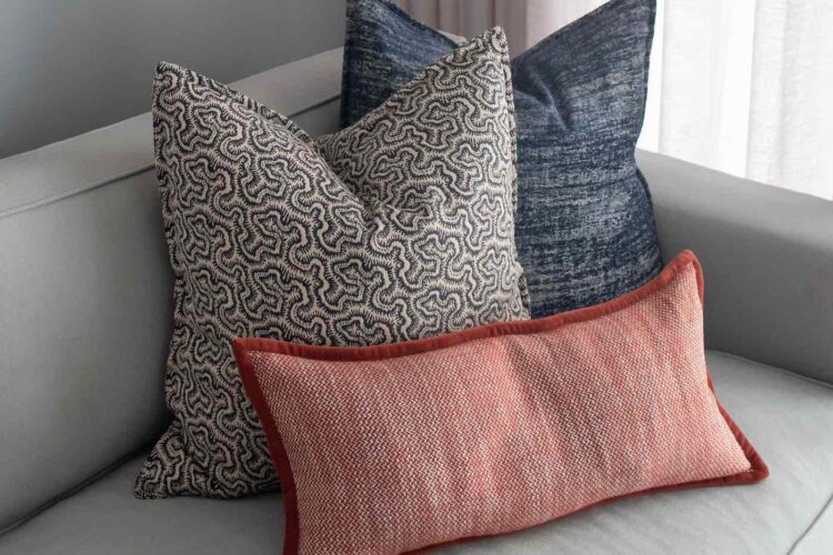 Three decorative pillows in various textures and colors rest on a light gray sofa. The background features soft fabric drapes, enhancing the cozy living space ambiance.