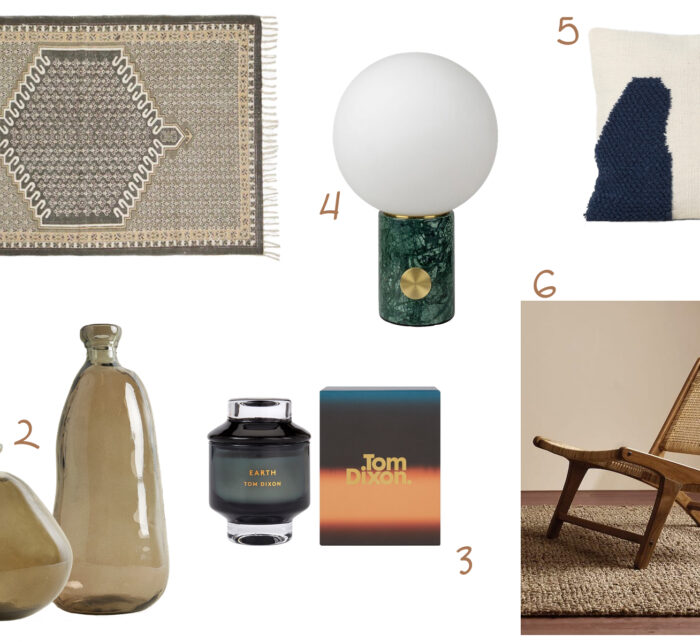 A decorative rug lies flat, two glass vases rest beside a candle in a black jar, a round lamp stands on a green marble base, and a textured throw pillow sits near a wooden chair on a woven mat.