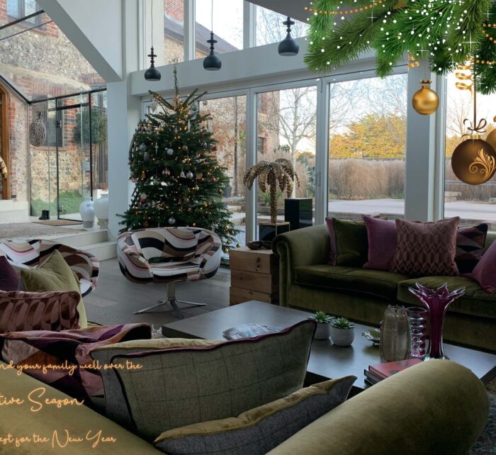 A beautifully decorated living room features a Christmas tree and a cozy seating area. Large glass windows display a scenic outdoor view, while hanging ornaments add a festive touch.