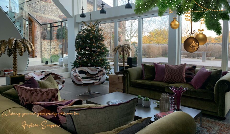 A beautifully decorated living room features a Christmas tree and a cozy seating area. Large glass windows display a scenic outdoor view, while hanging ornaments add a festive touch.