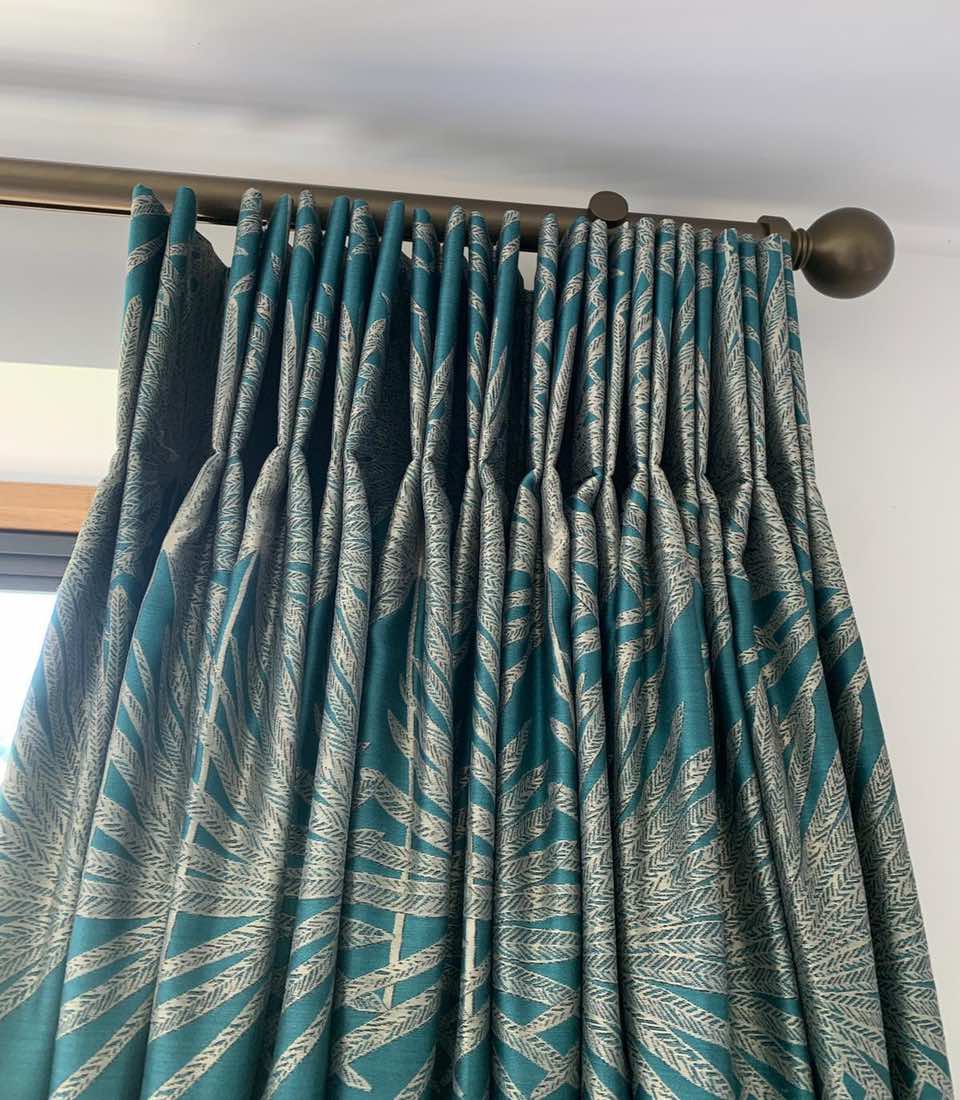 Turquoise and patterned curtains hang in folds from a bronze rod, partially covering a window, creating a decorative accent in a bright indoor space.