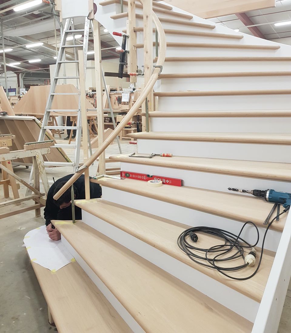 A person kneels on the wooden steps of a spiral staircase, working with paper and tools. The scene is set in a workshop, with other furniture pieces in the background.