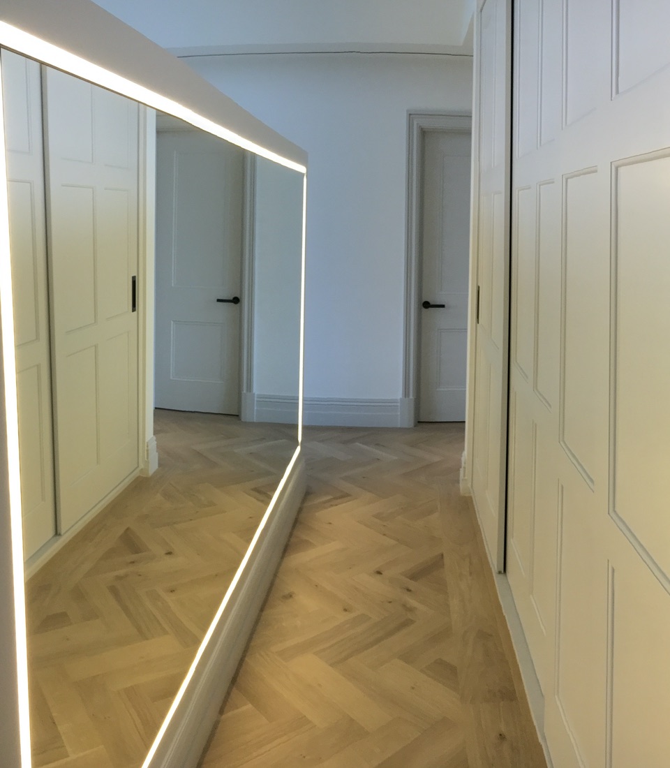 A rectangular, backlit mirror reflects a spacious, brightly lit hallway with light-colored herringbone flooring. White walls and doors create a clean, modern aesthetic, enhancing the sense of openness.