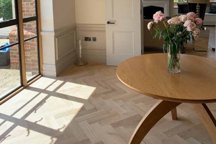 A round wooden table with a vase of pink flowers sits on a light wood herringbone floor. Sunlight filters through large windows, illuminating the modern interior with a door leading to another room.