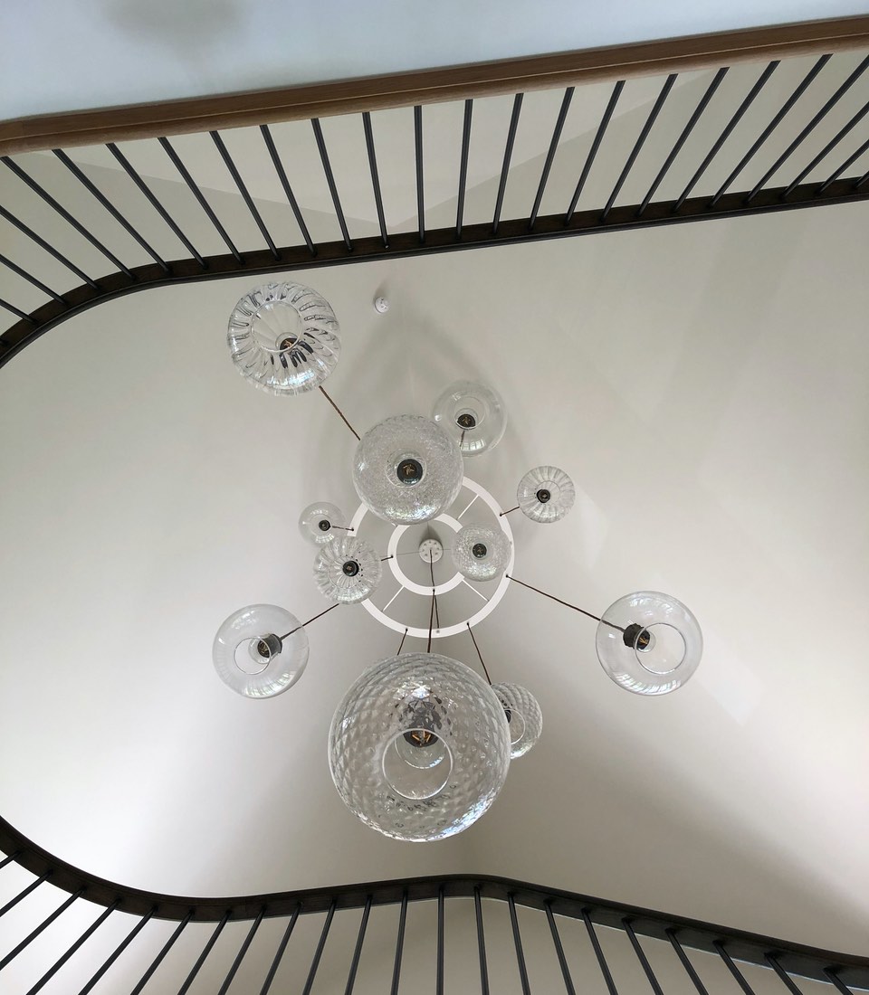 A modern chandelier composed of various glass globes hangs from a circular fixture, illuminating a curved stairway with black railings and white walls, creating an elegant interior atmosphere.