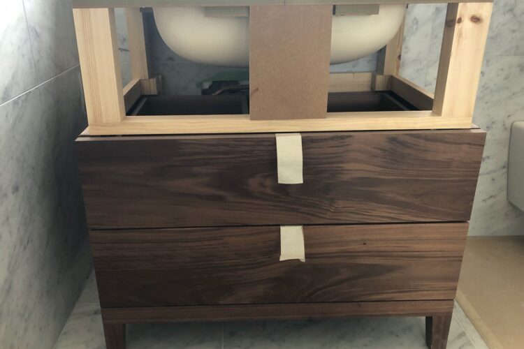 A wooden vanity with two drawers and a light wood frame holds a white basin above it. The backdrop features marble walls, indicating a bathroom setting under natural light.
