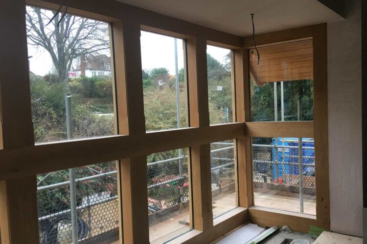 Wooden-framed windows are installed in a room, allowing a view of trees and a construction area outside. The floor is unfinished, with tools and materials scattered nearby.