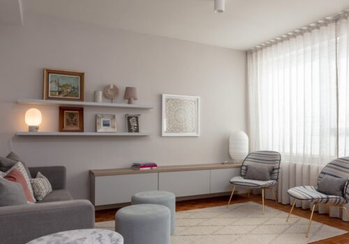 A modern living room features a gray sofa with decorative pillows, two patterned chairs, circular tables, and a low cabinet. Shelves display framed art and a lamp, while sunlight filters through sheer curtains.