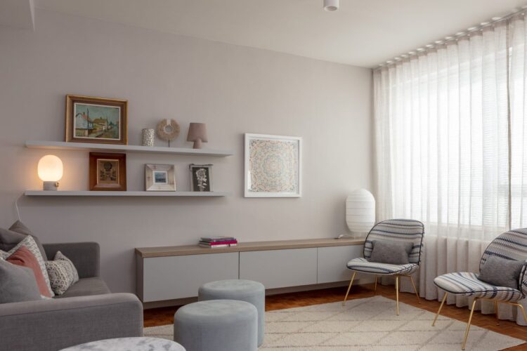 A modern living room features a gray sofa with decorative pillows, two patterned chairs, circular tables, and a low cabinet. Shelves display framed art and a lamp, while sunlight filters through sheer curtains.