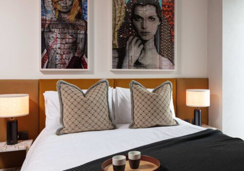 A stylish bed with decorative pillows is centered in a modern room. Above, two framed art pieces add color. A side table holds a tray with cups, complemented by ambient lamps.