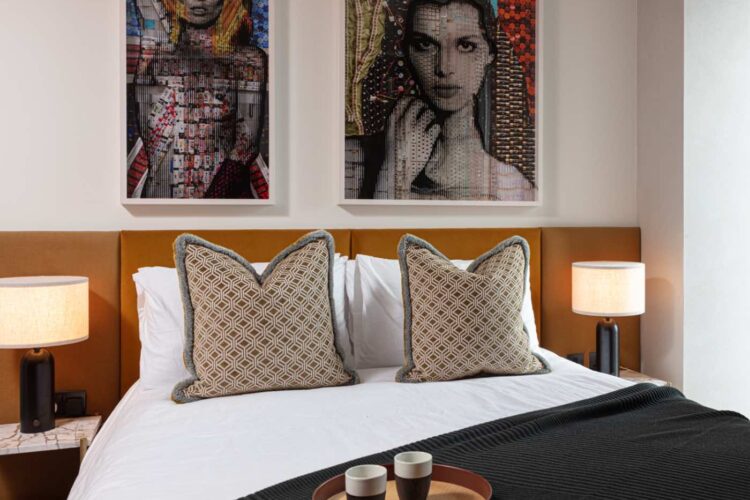 A stylish bed with decorative pillows is centered in a modern room. Above, two framed art pieces add color. A side table holds a tray with cups, complemented by ambient lamps.
