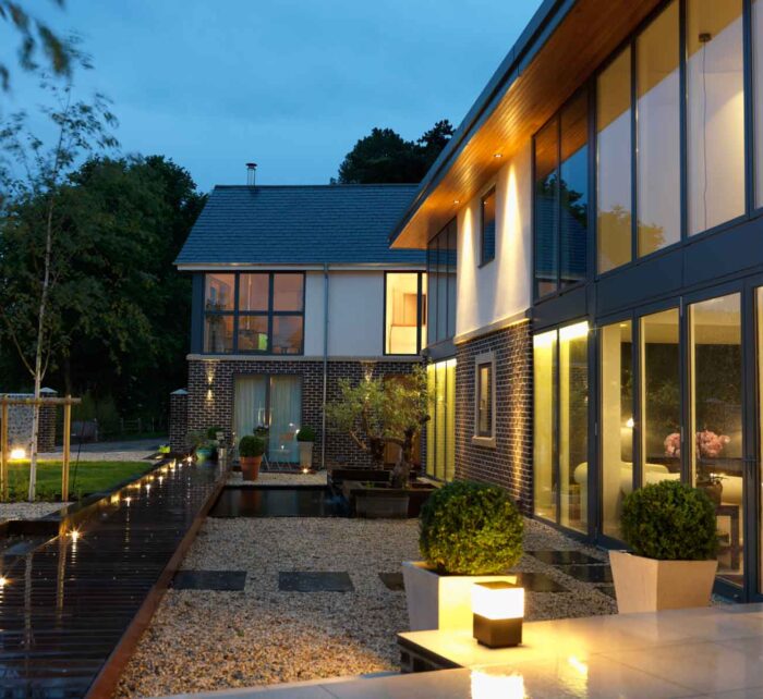 A modern house with large windows reflects warm lights in the evening. A wooden pathway leads through a landscaped garden with pebbles, planters, and subtle illumination, surrounded by trees and greenery.