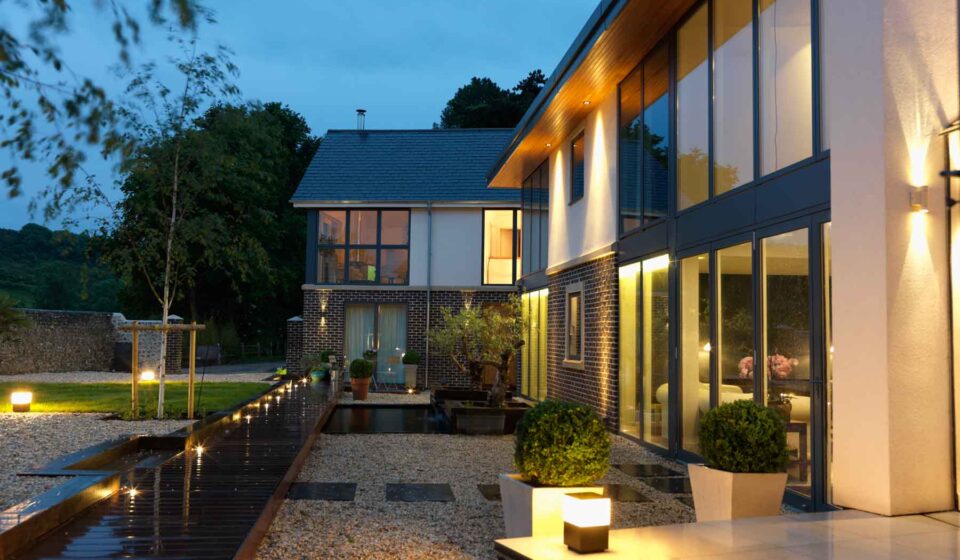 A modern house with large windows reflects warm lights in the evening. A wooden pathway leads through a landscaped garden with pebbles, planters, and subtle illumination, surrounded by trees and greenery.