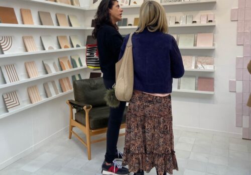 Two women are engaged in conversation in a gallery-like space, surrounded by shelves displaying various art pieces or notebooks, with a low wooden chair nearby.