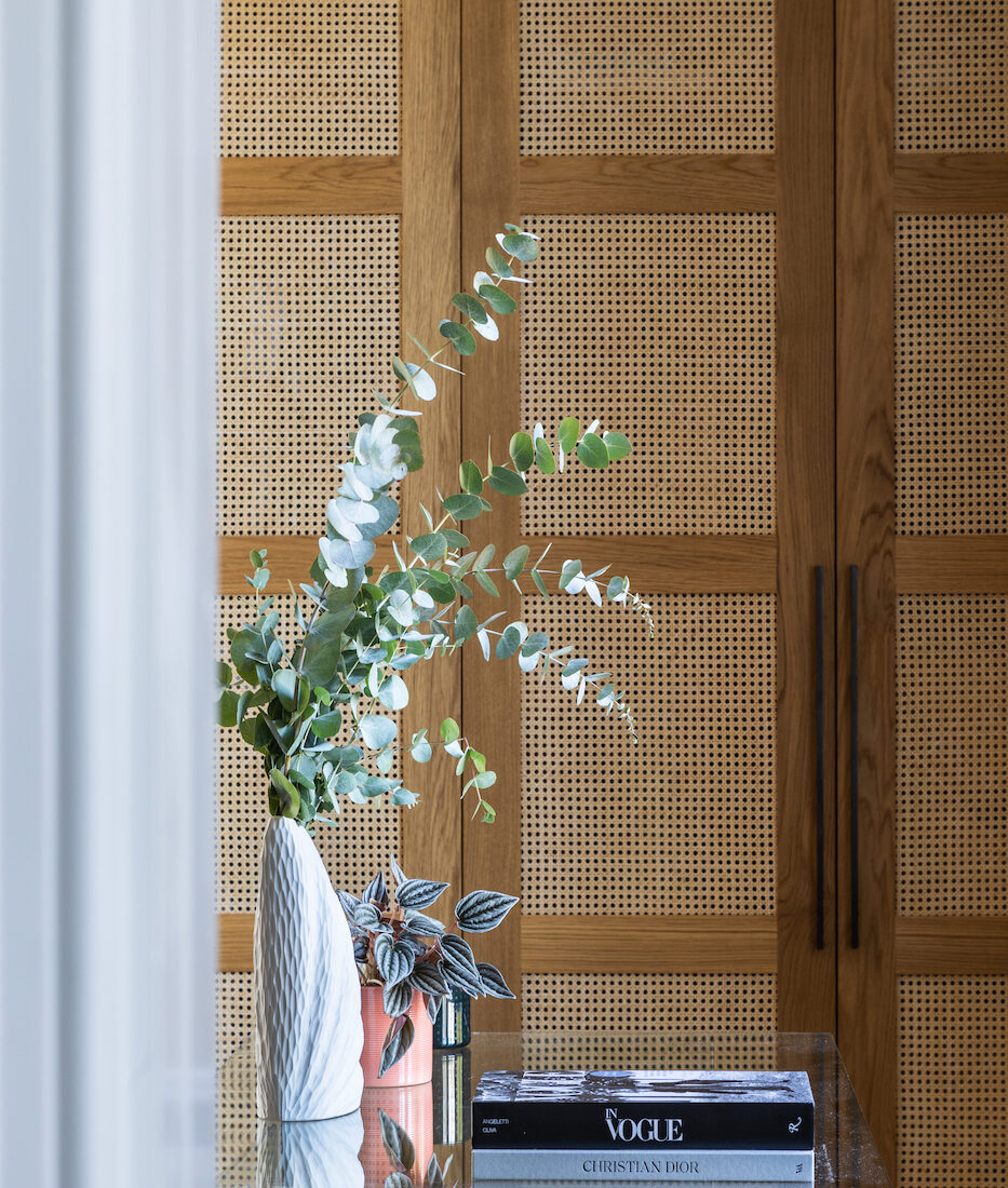A white vase with green eucalyptus stands on a glass table. Nearby, books with titles like "In Vogue" and "Christian Dior" rest, against a backdrop of wooden, perforated doors.