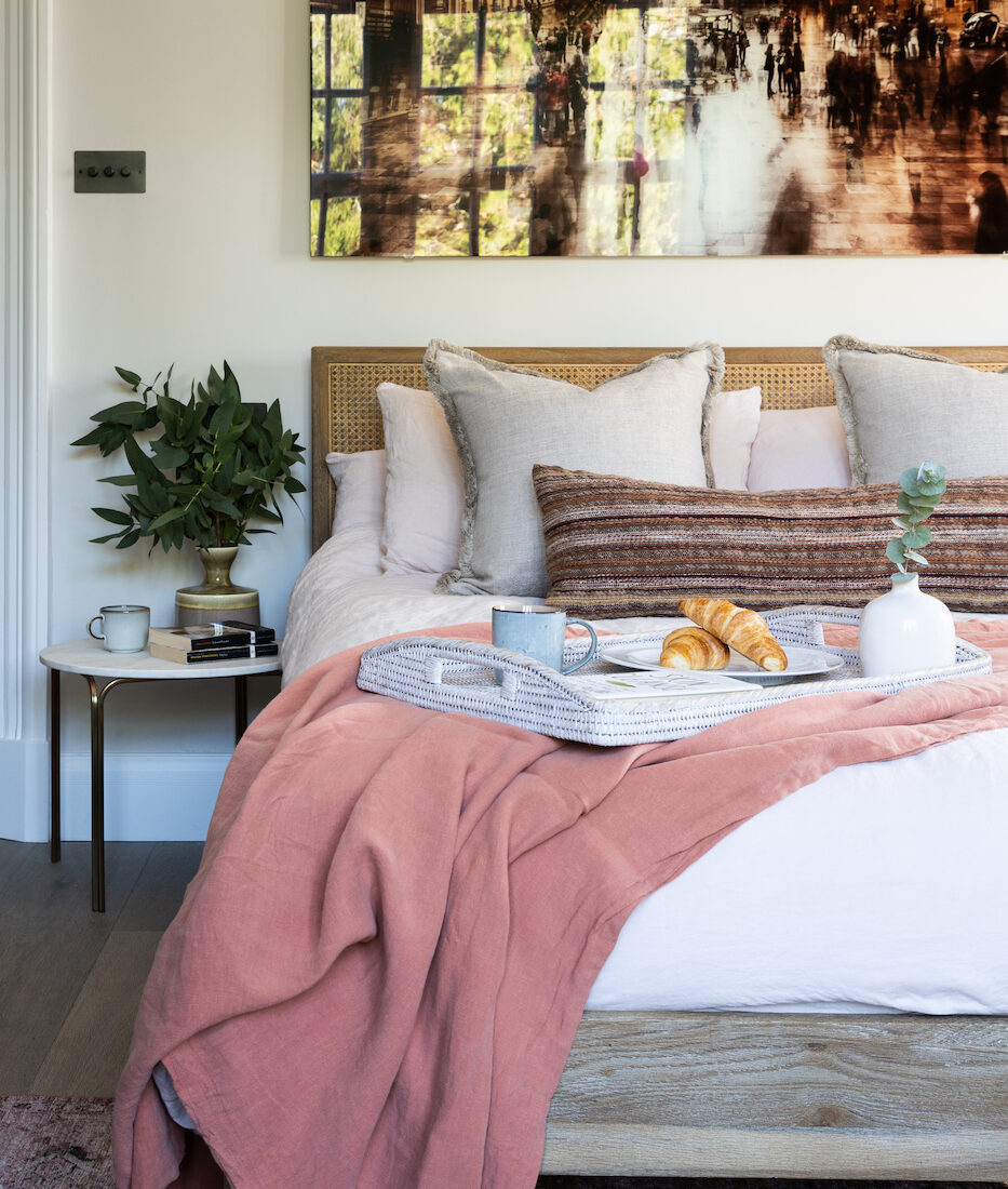 A bed with decorative pillows is the focal point, featuring a tray with croissants and a vase. The room is bright, with a plant nearby and an artistic photo on the wall.