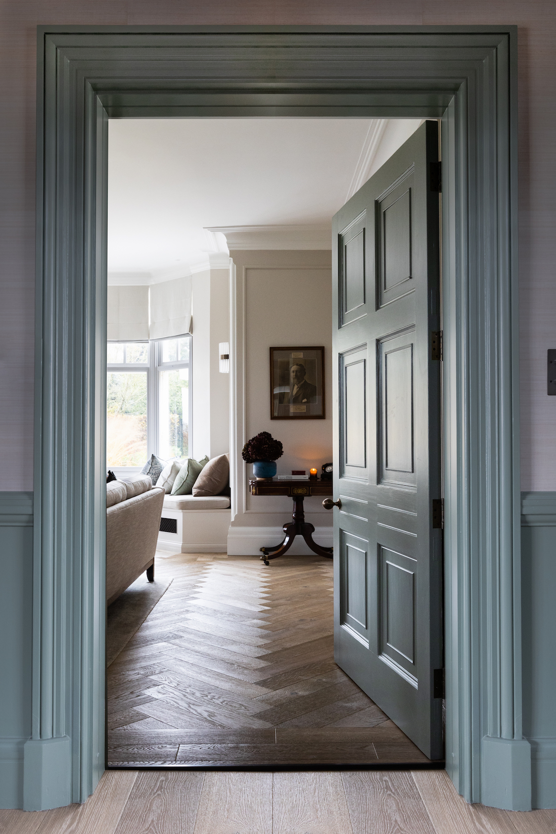 A partially open green door reveals a cozy room with a beige sofa, decorative pillows, and an elegant table with a candle, amid a lightly textured wooden floor.